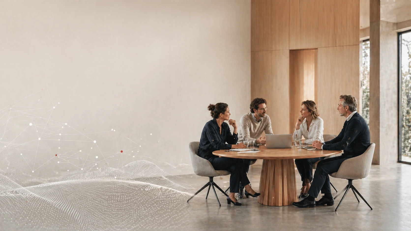 Four people sit around a round wooden table in a modern office, engaged in discussion with a laptop open.