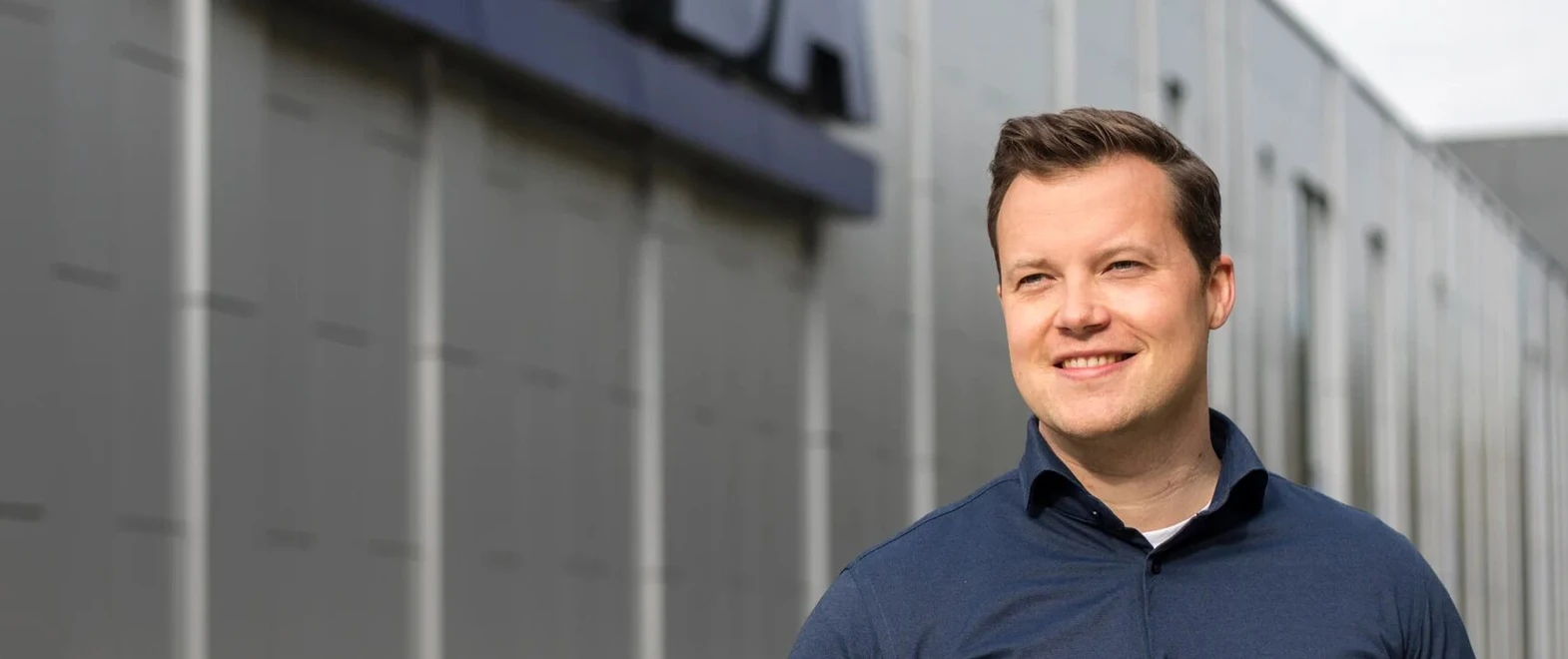 Smiling man in a blue shirt stands outdoors in front of a modern gray building with a blurred logo.