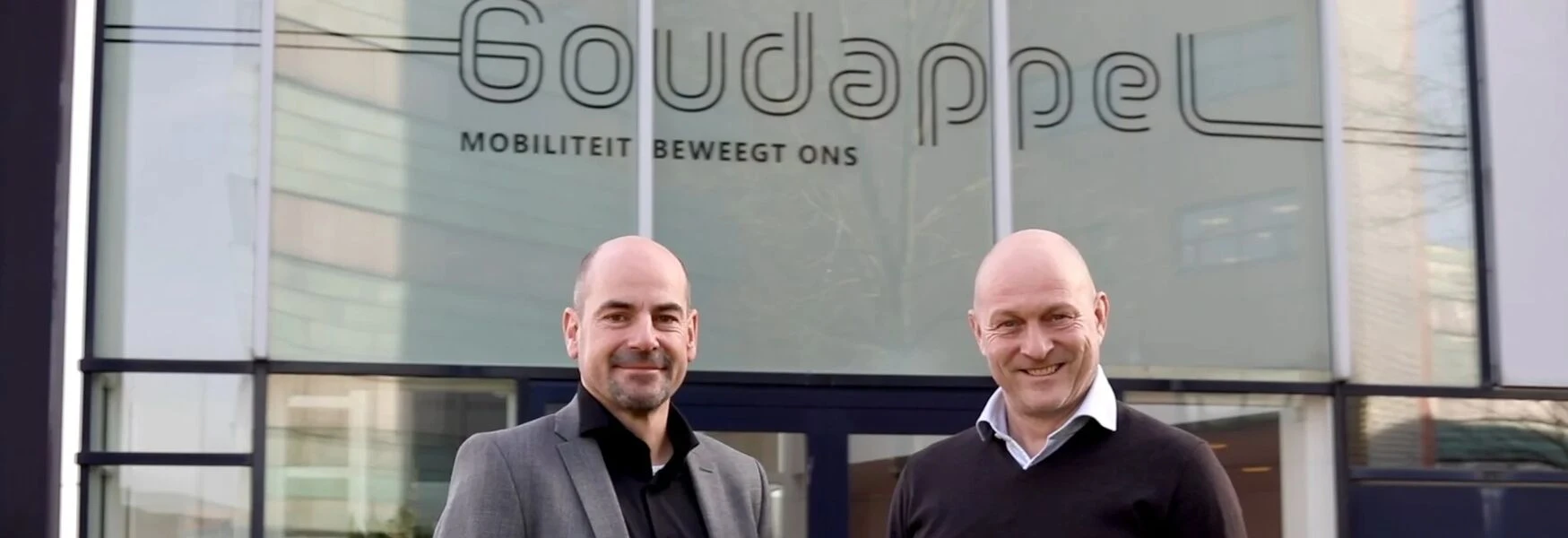 Two men standing in front of a building with the sign "Goudappel Mobiliteit Beweegt Ons."