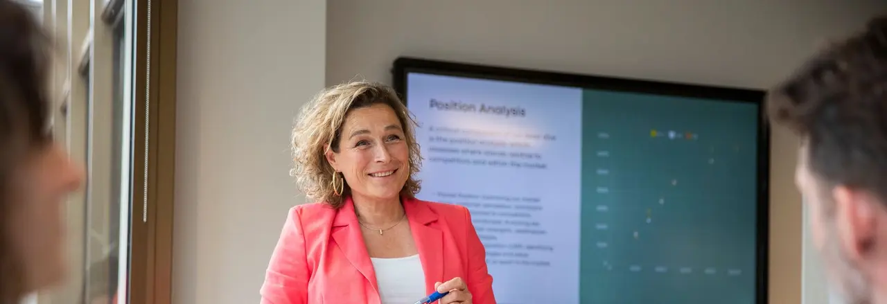 Woman in a coral blazer presents a position analysis on a screen to two people in a meeting room.