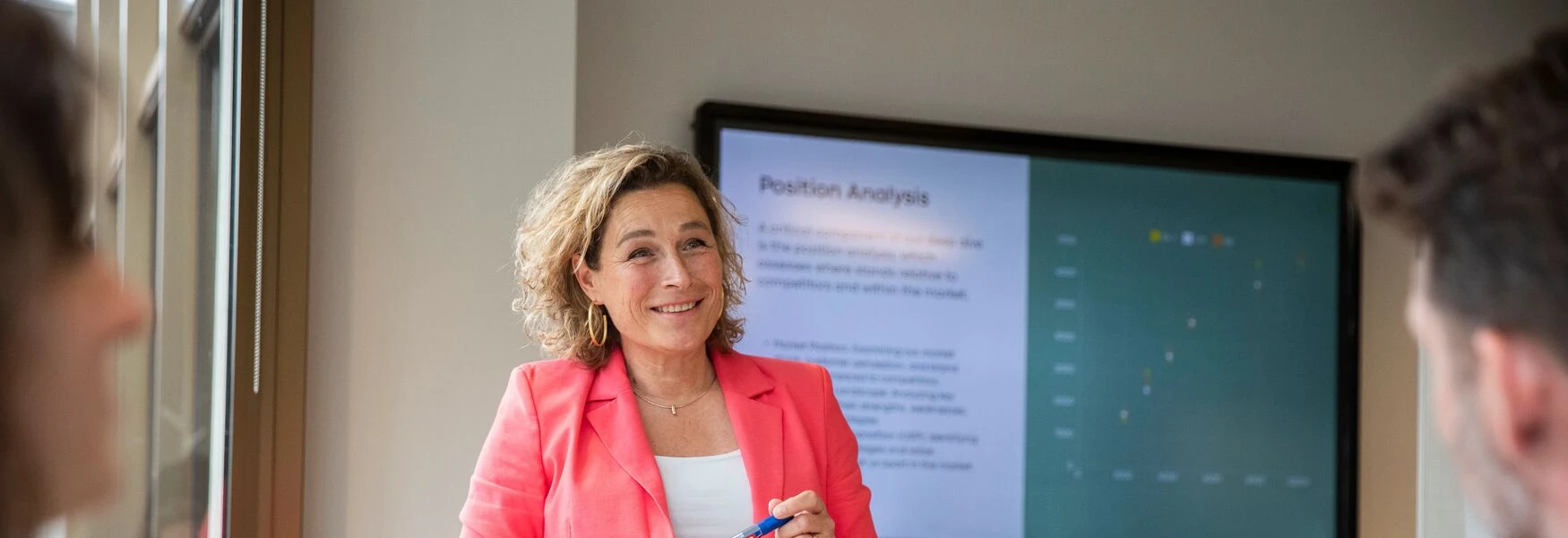 Woman in a coral blazer presents a position analysis on a screen to two people in a meeting room.