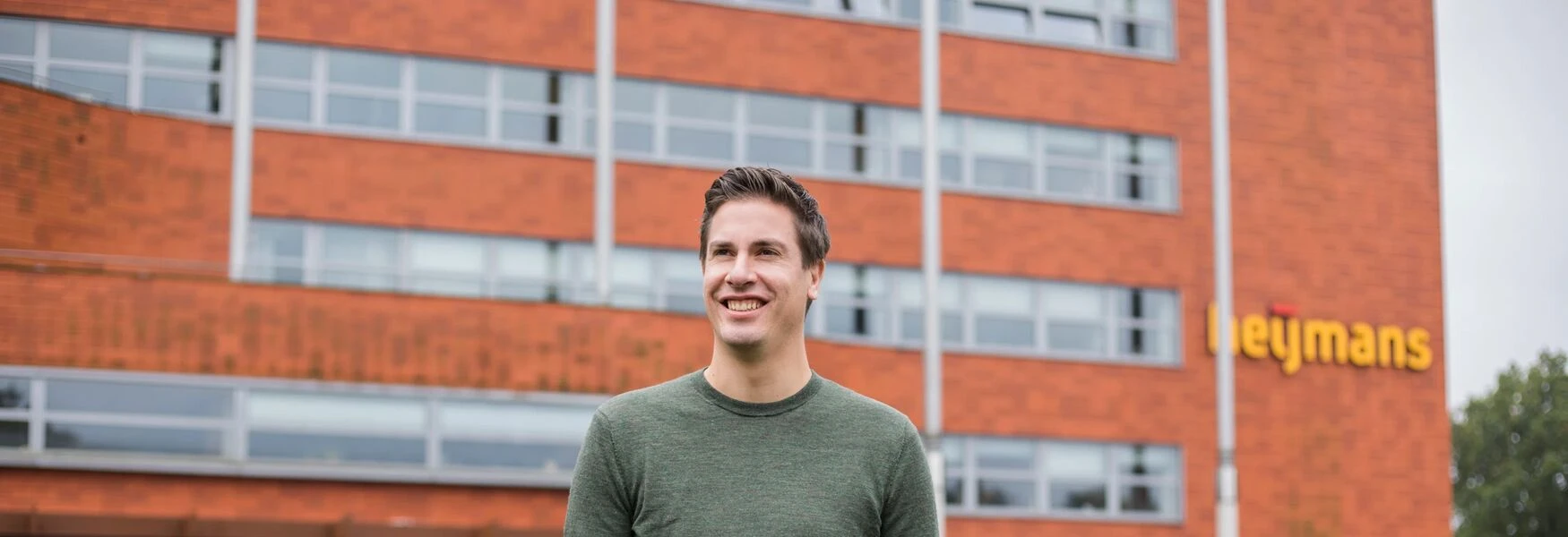A person in a green sweater stands smiling in front of a brick building with large windows and the word "Heijmans" visible.