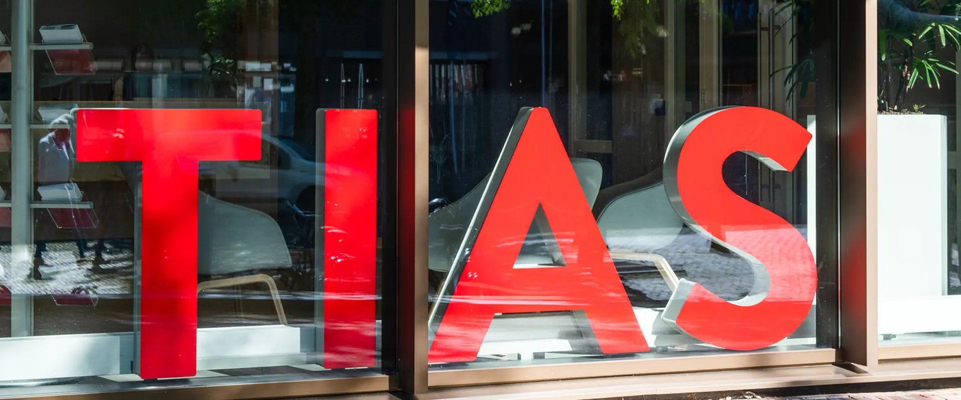 Large red letters spelling "TIAS" are displayed on a glass window, with reflections of the street and indoor plants visible.