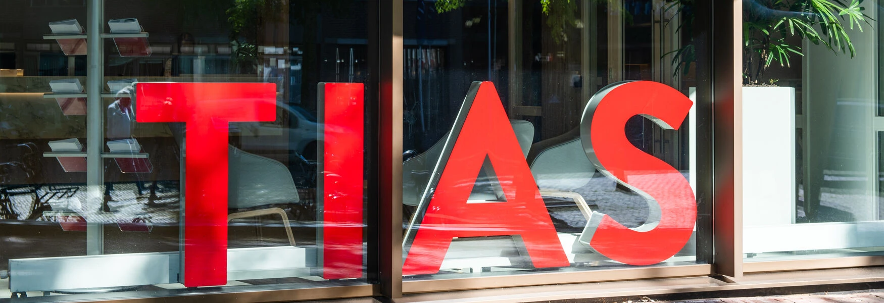 Large red letters spelling "TIAS" are displayed on a glass window, with reflections of the street and indoor plants visible.