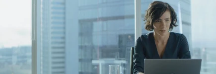 A woman with short hair works on a laptop in a modern office with large windows and cityscape views.