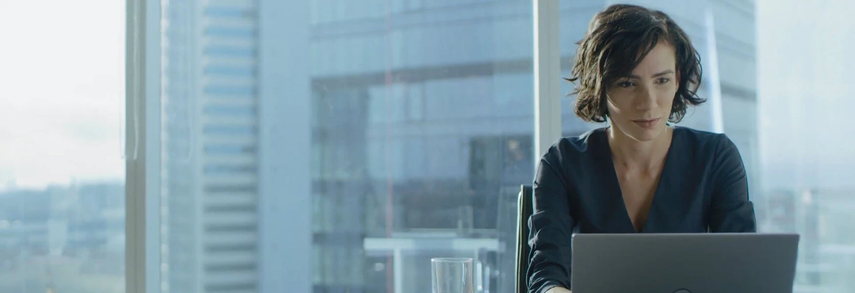 A woman with short hair works on a laptop in a modern office with large windows and cityscape views.