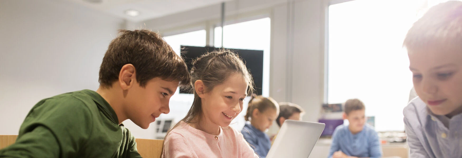 Children engaged in learning, gathered around a laptop in a bright classroom setting.