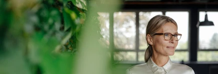 A woman with glasses, wearing a light blouse, stands thoughtfully near a window with greenery in the foreground.