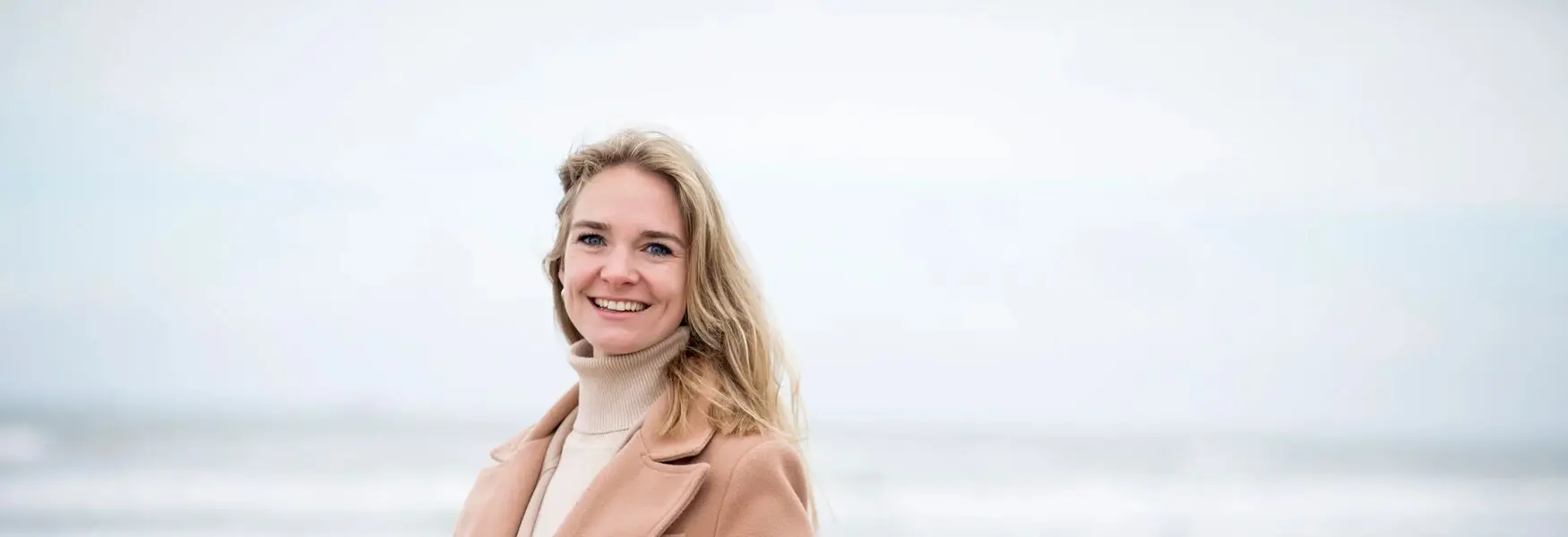 Smiling person in a beige coat stands outdoors with a blurred background.