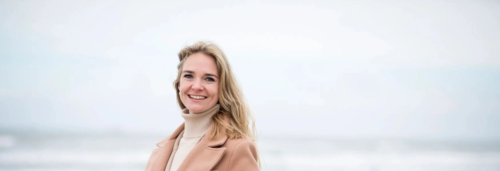 Smiling person in a beige coat stands outdoors with a blurred background.