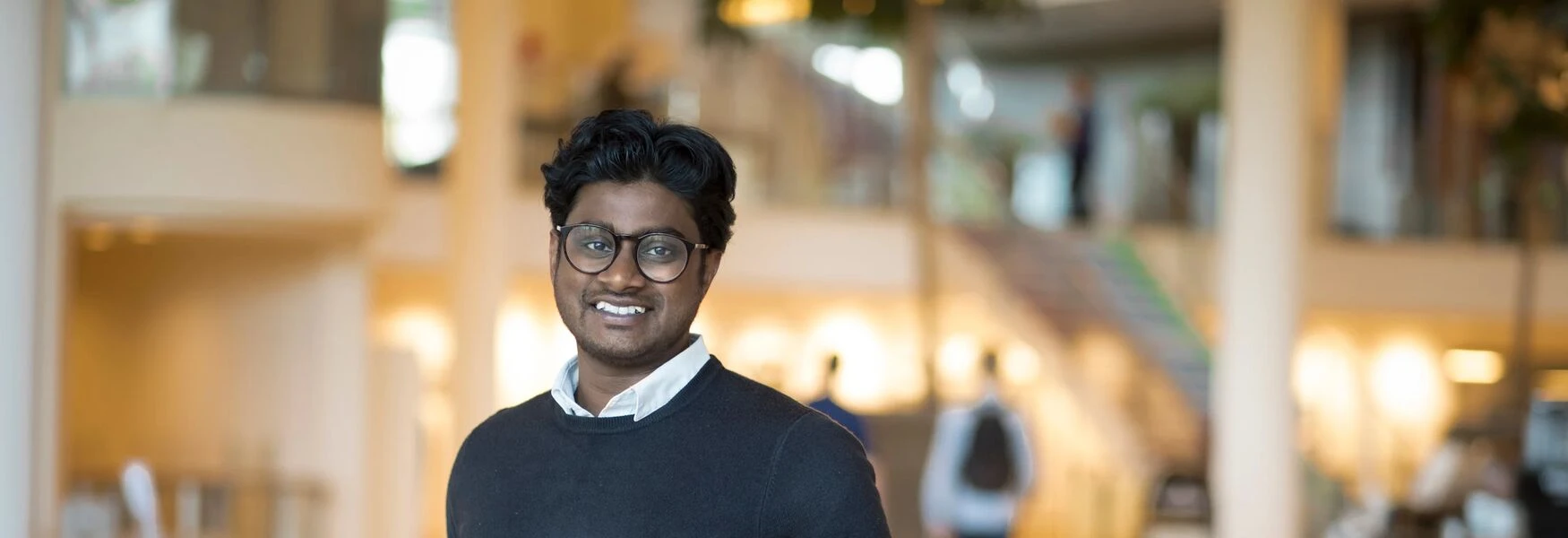 Person smiling indoors, wearing glasses and a sweater, with a blurred background of a spacious, well-lit room.
