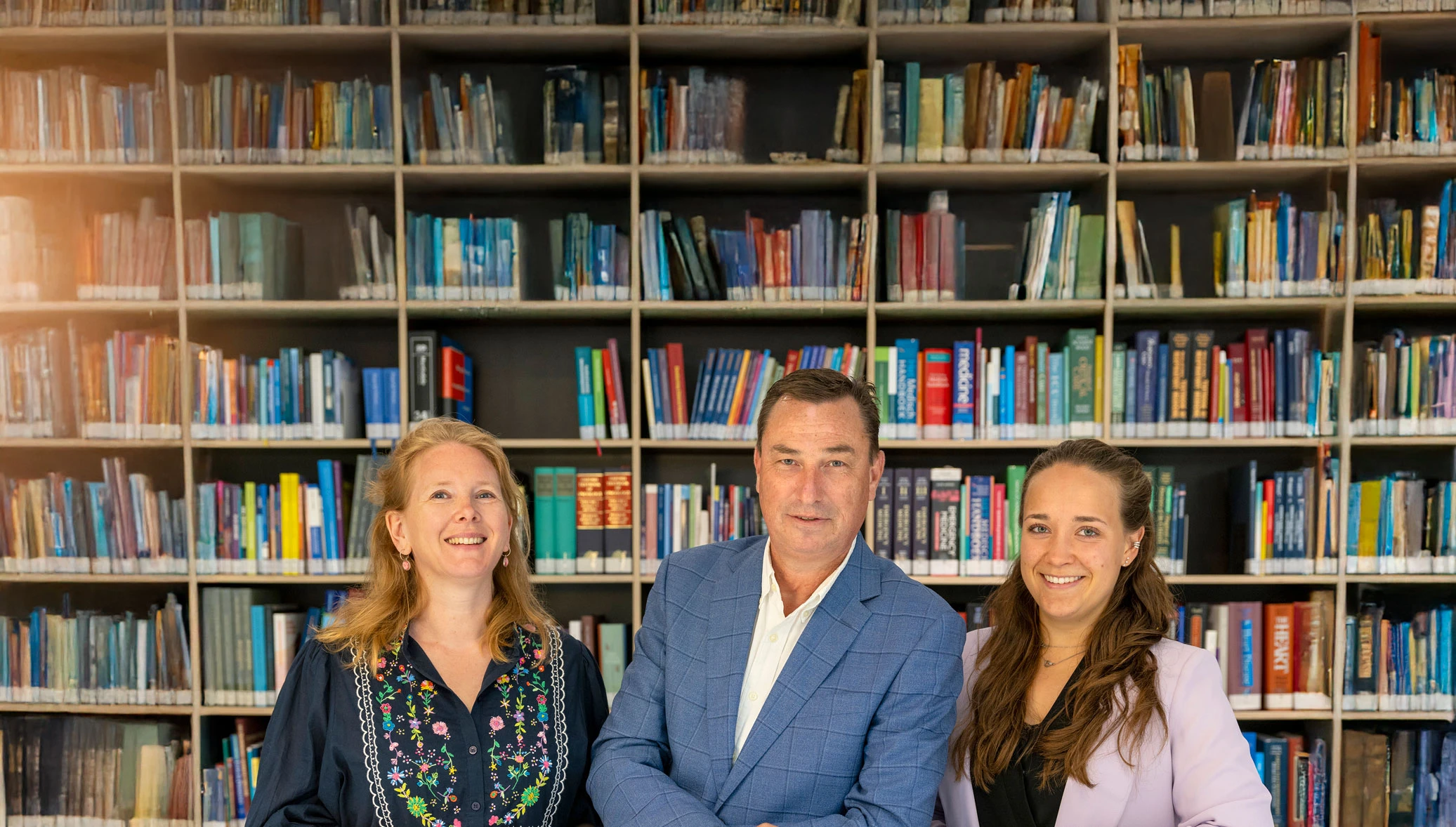 Three people smiling in front of a large bookshelf filled with colorful books.