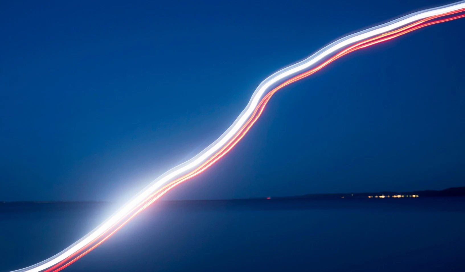 Bright, curving light streaks against a deep blue evening sky and calm water in the background.