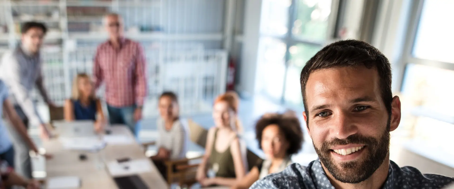 A man takes a selfie with a group of people sitting around a table in a bright office setting.