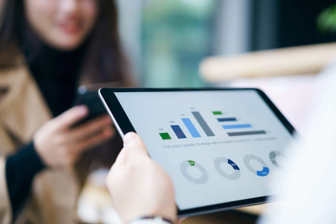 Person holding a tablet displaying bar and pie charts, while another person uses a smartphone in the background.