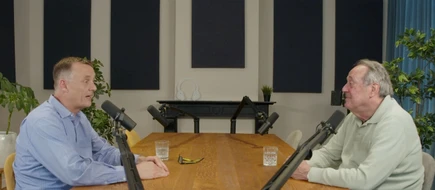 Two men sitting across a wooden table in a podcast studio, with microphones and water glasses in front of them.