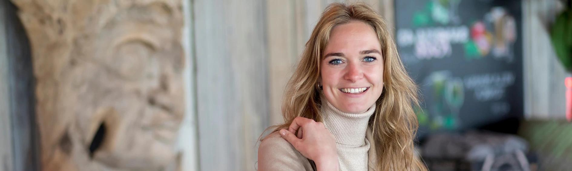 Smiling woman with long blonde hair, wearing a beige turtleneck, stands indoors with a blurred background featuring artwork and greenery.