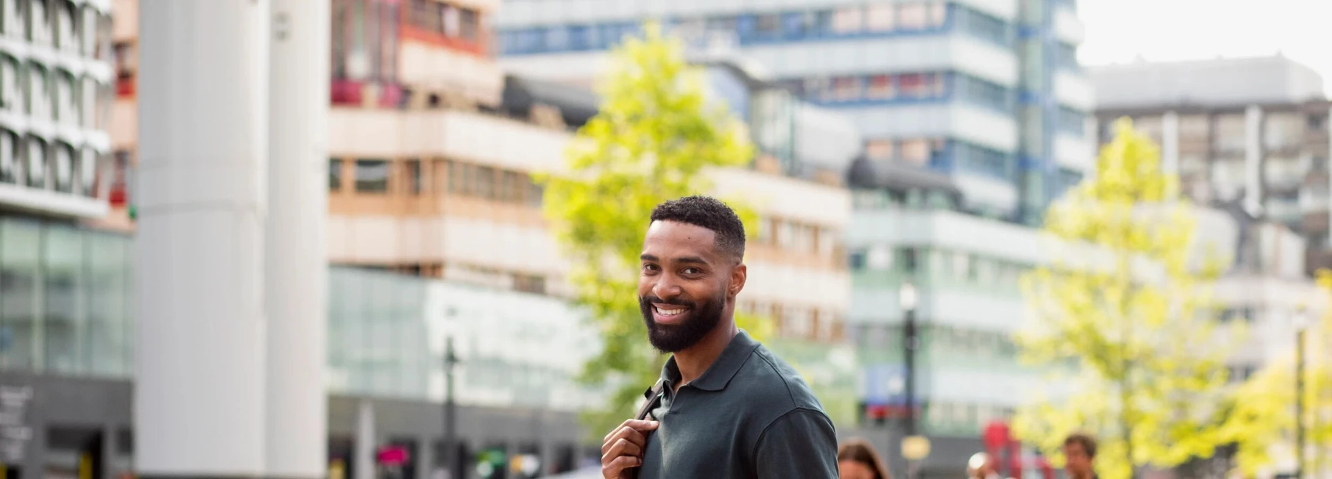 Smiling man with a backpack stands in an urban setting, with modern buildings and trees in the background.