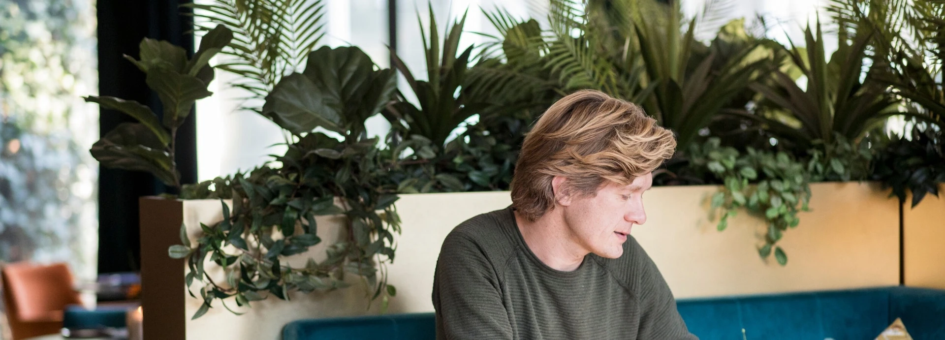 A person with blonde hair sits on a blue couch in a modern setting, surrounded by lush green plants.