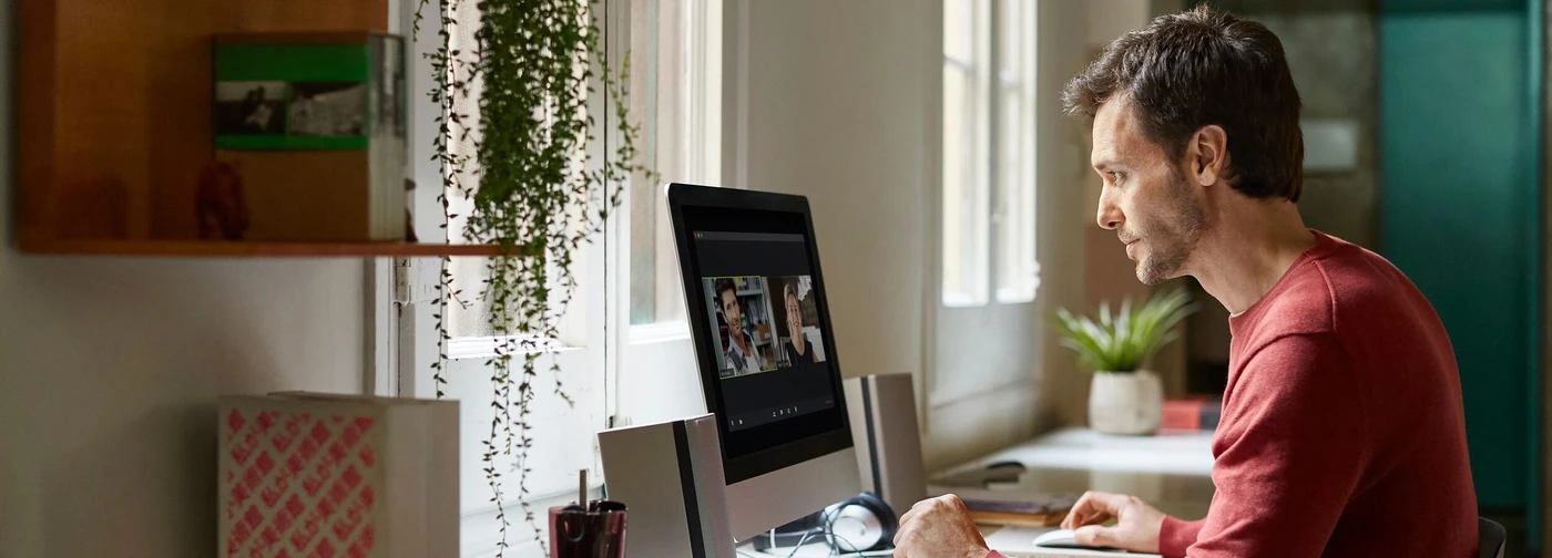 Man in a red shirt working on a computer at a desk near a window, with a plant and office supplies nearby.