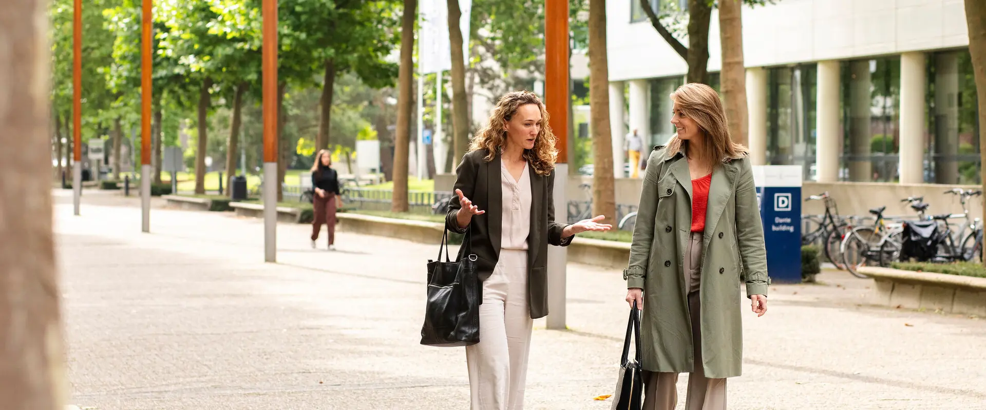 Two women walking and talking on a tree-lined path along a building with bicycles parked nearby.