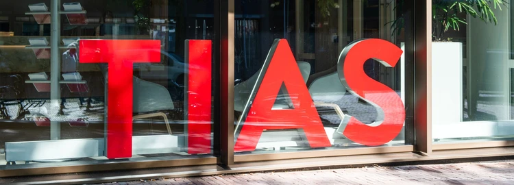 Large red letters spelling "TIAS" displayed in a window, reflecting a street scene with trees and parked cars.