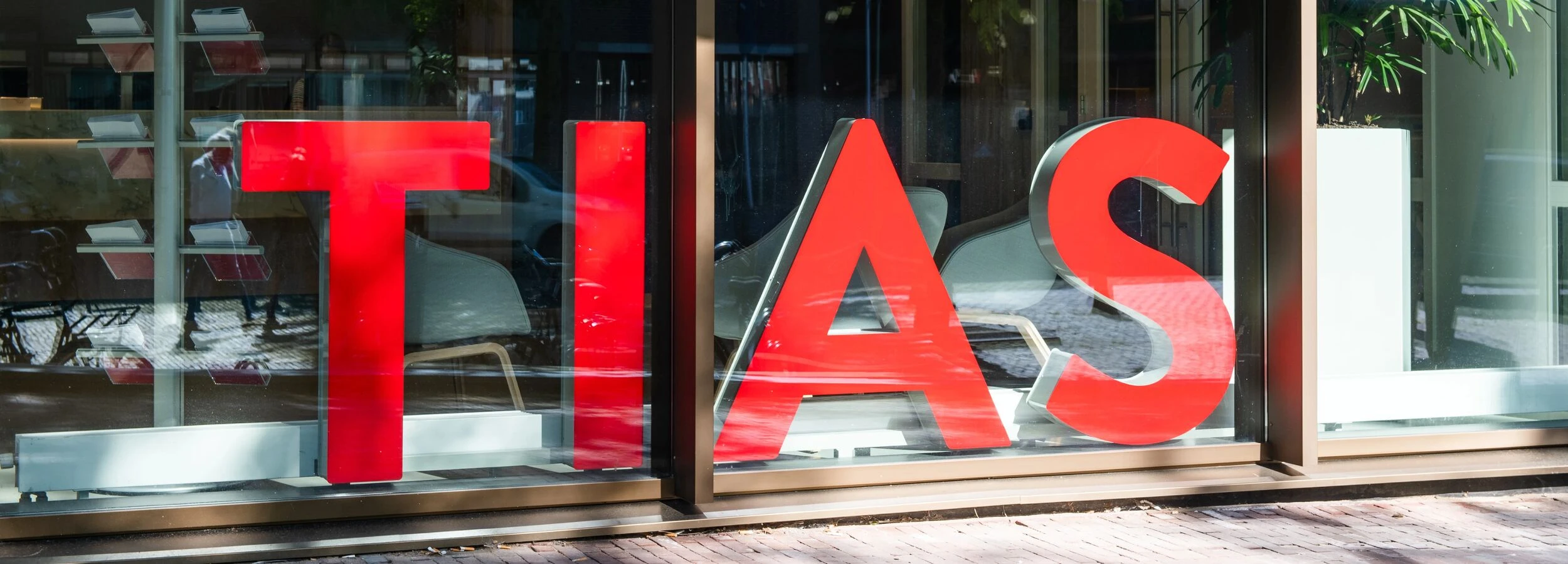 Large red letters spelling "TIAS" displayed in a window, reflecting a street scene with trees and parked cars.