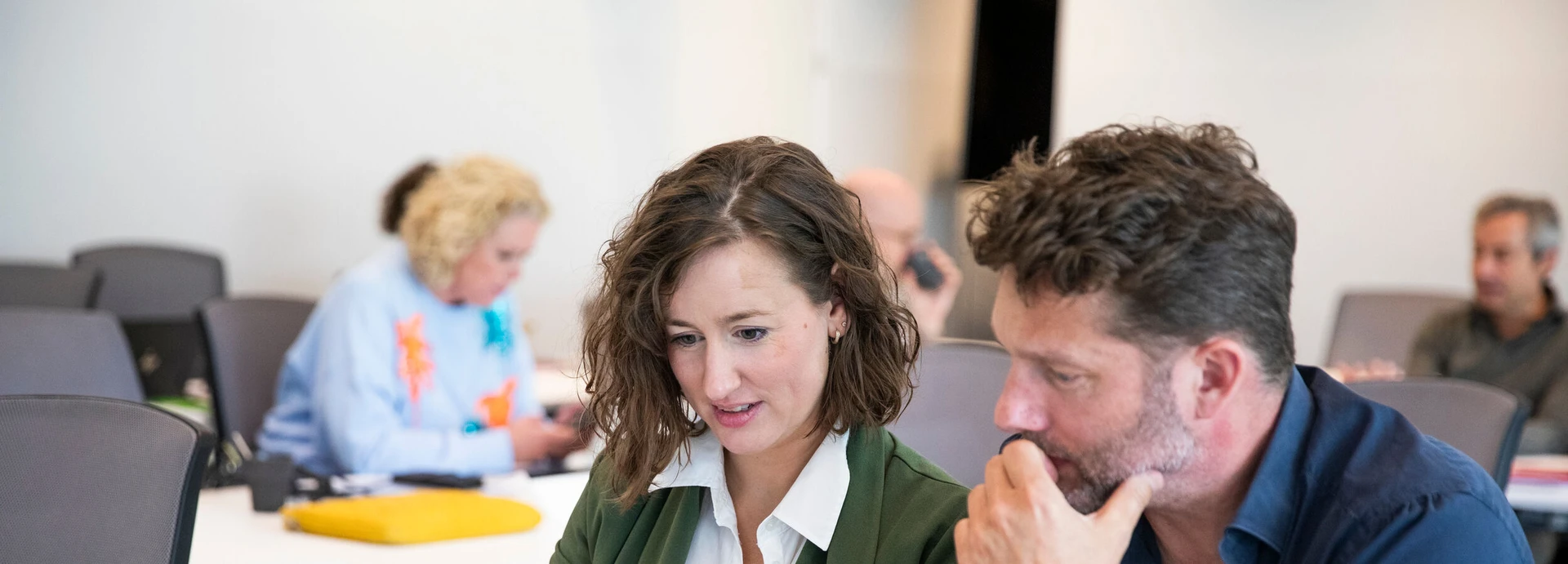 Two people engaged in discussion at a conference table, with others working in the background.