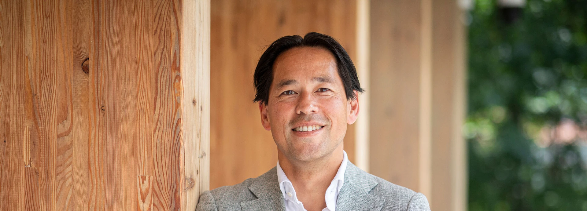 Smiling man in a gray blazer and white shirt stands next to a wooden wall with greenery in the background.