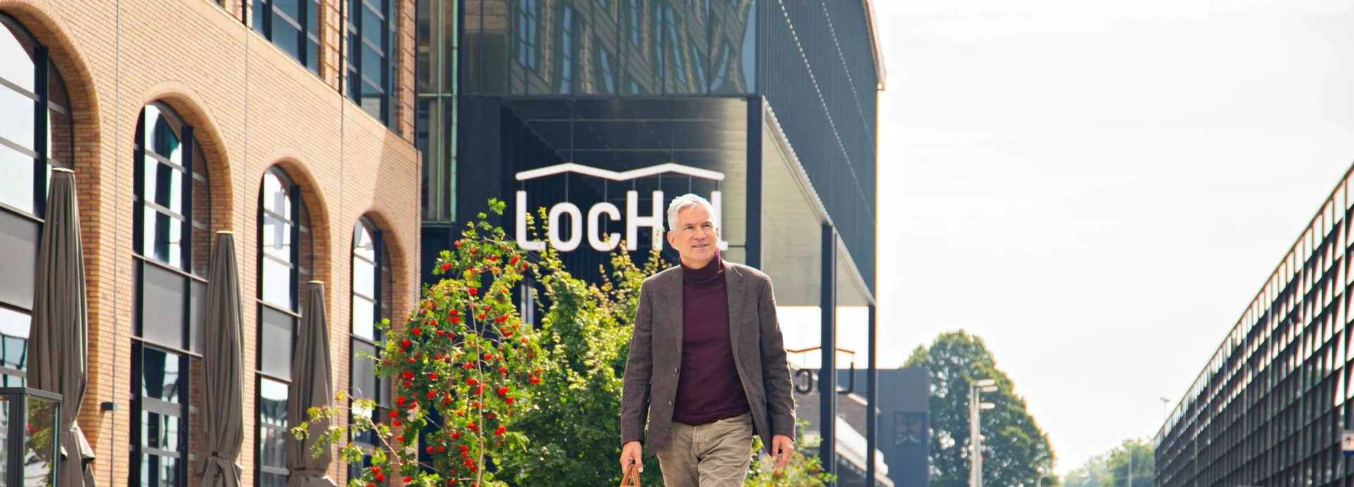 Man walking outside a modern building with "LoCH" sign, surrounded by greenery and clear sky.