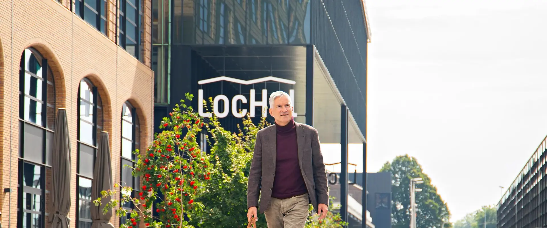 Man walking outside a modern building with "LoCH" sign, surrounded by greenery and clear sky.