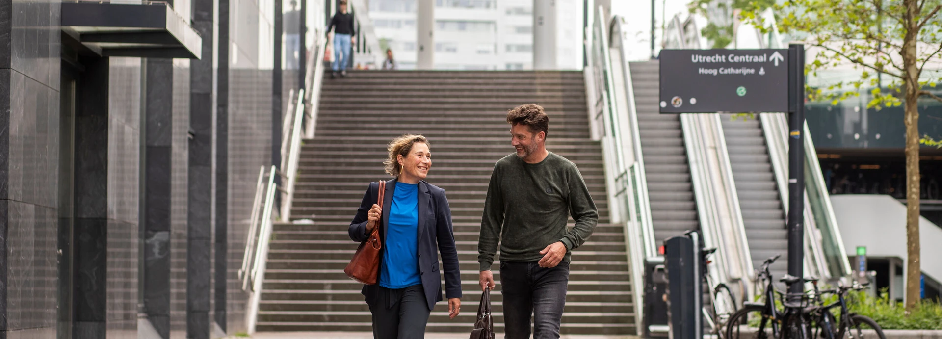 Two people walking and talking near a staircase at Utrecht Central, with a bicycle rack and a sign visible in the background.