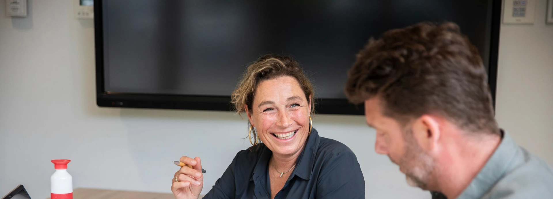 A woman smiles while holding a pen, sitting at a table with a man. A large screen is visible in the background.