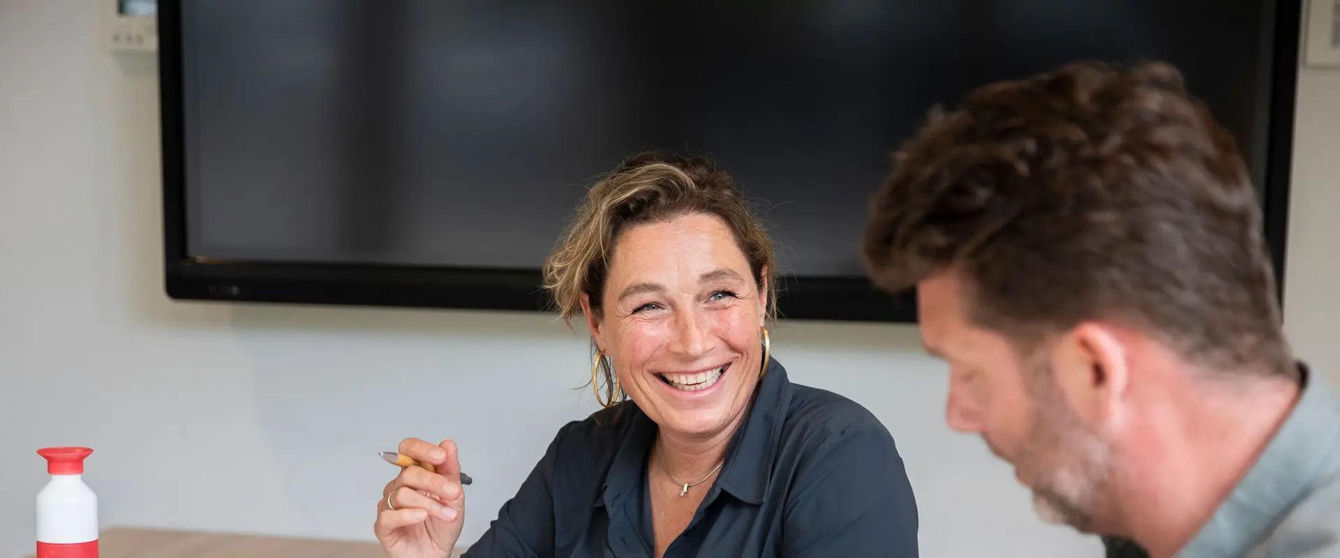 A woman smiles while holding a pen, sitting at a table with a man. A large screen is visible in the background.