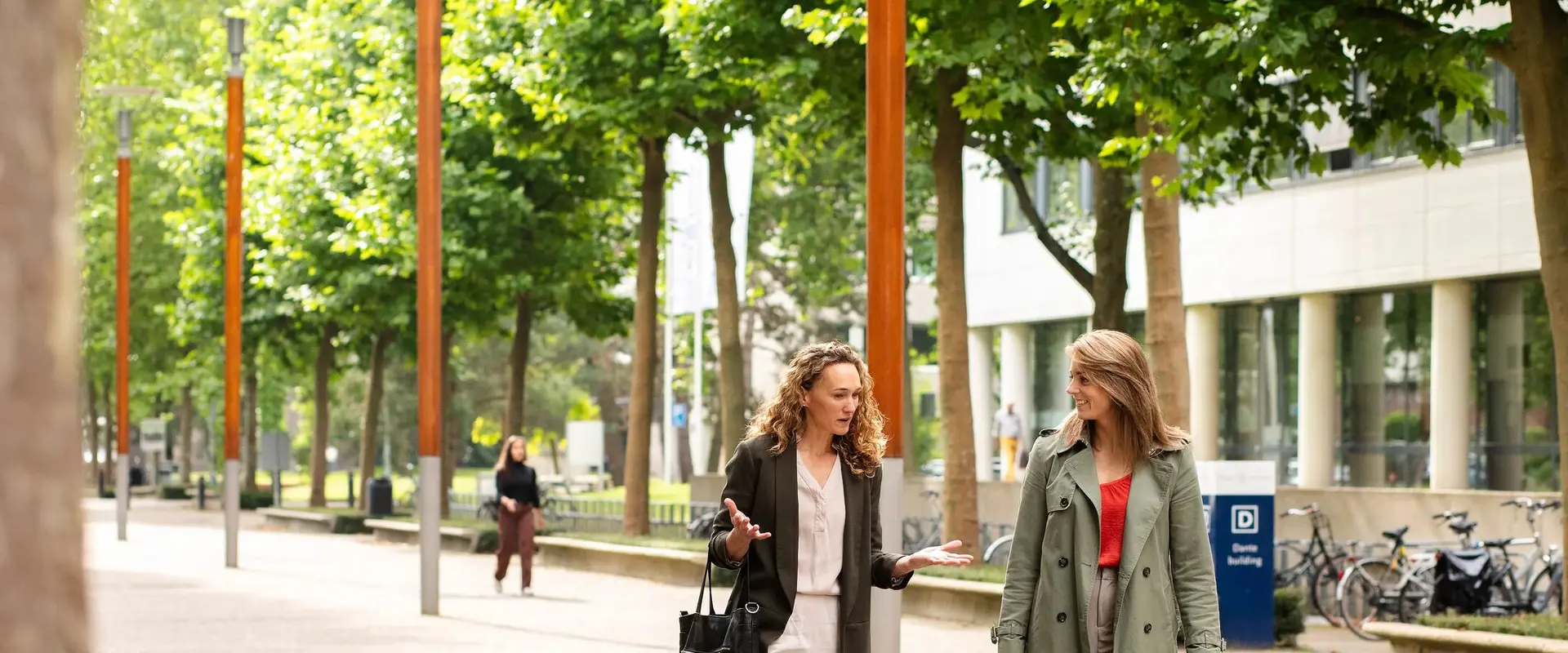 Two people walking and talking on a tree-lined path outside a modern building, with bicycles parked nearby.