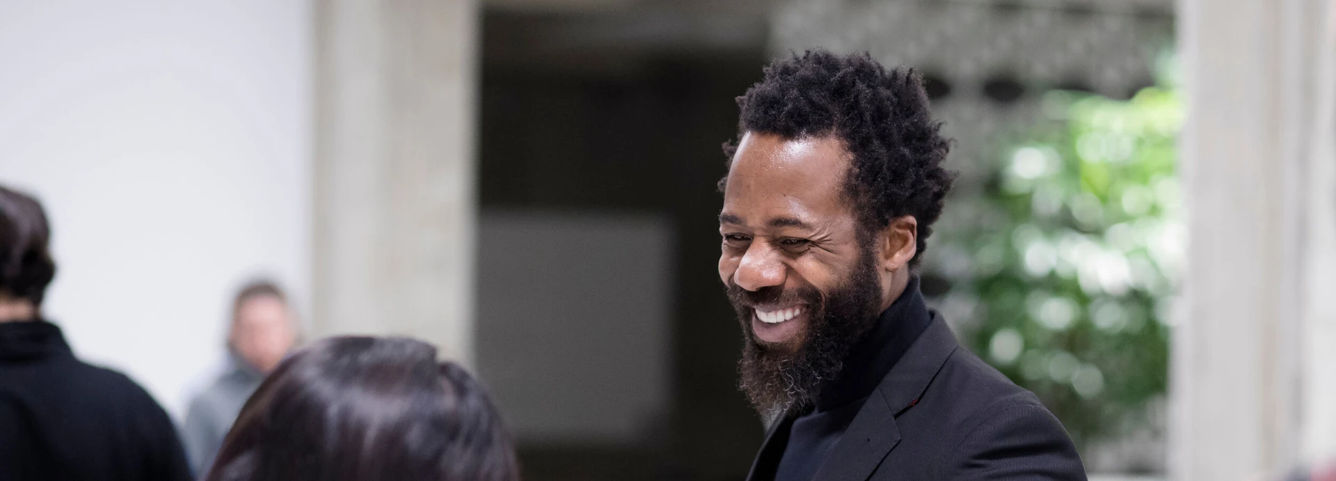 A man with a beard and curly hair smiling while talking to someone, standing indoors with a blurred background.