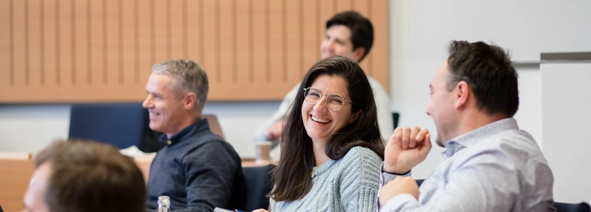 People sitting in a classroom, smiling and engaged in conversation, with a woman in glasses at the center.