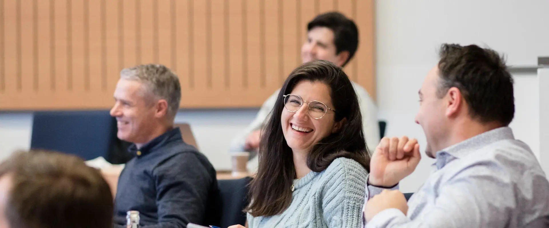 People sitting in a classroom, smiling and engaged in conversation, with a woman in glasses at the center.