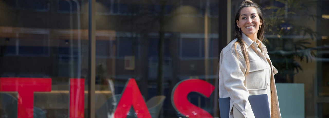 A woman smiling while holding a laptop, standing in front of a building with large red letters.