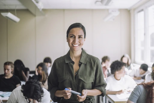 Smiling teacher in a classroom stands holding a pen, with students seated and working at desks in the background.