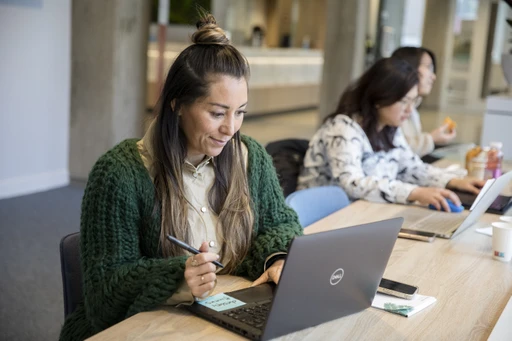 A woman with long hair works on a laptop at a wooden table in an office, with two other people focused on their laptops in the background.