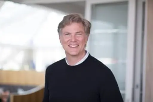 Smiling man in a black sweater standing in a modern office setting with blurred background.