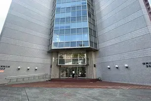 Entrance of a modern building with large glass windows, a wide staircase, and a brick facade. Two people are near the doorway.