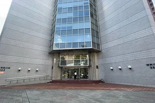 Entrance of a modern building with large glass windows, a wide staircase, and a brick facade. Two people are near the doorway.