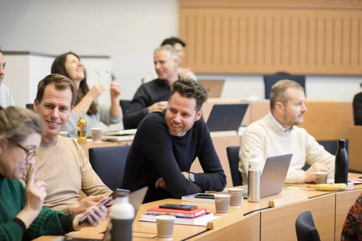 Students engaging in discussion and using laptops and phones in a classroom setting with desks and chairs.