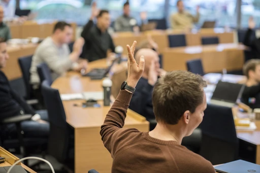 A classroom with students seated at desks, one student in the foreground raising a hand, engaging in a discussion.