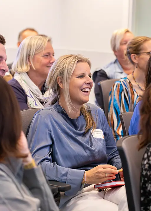 Smiling woman in a blue blouse sits among an engaged audience during a presentation or meeting.