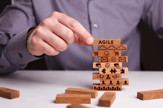 Person stacking wooden blocks labeled with symbols and "AGILE" on top, representing agile methodology concepts.