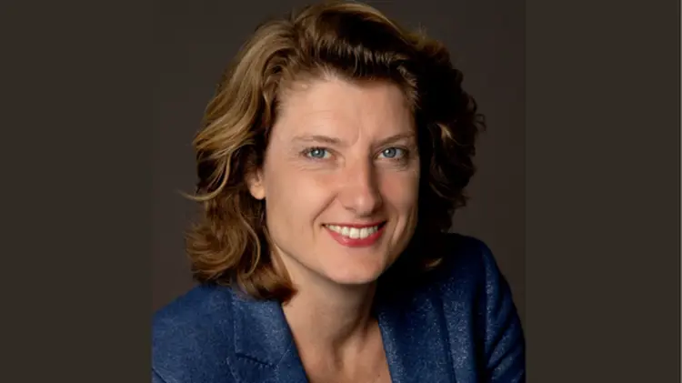 Portrait of a smiling woman with wavy light brown hair, wearing a blue blazer, set against a dark background.