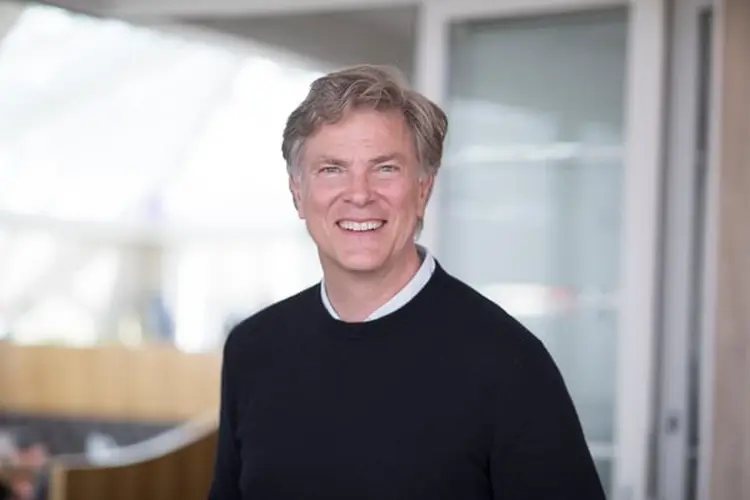 Smiling man in a black sweater stands indoors with a blurred background of modern architecture.